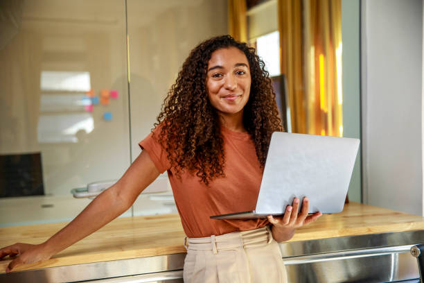 A smiling woman with curly hair holding a laptop in a modern office.