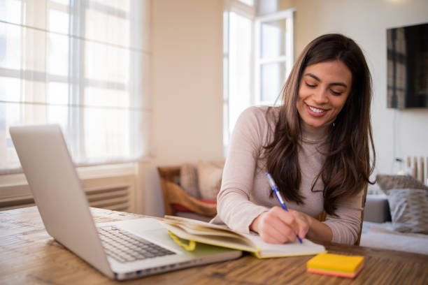 A woman smiling while taking notes in a notebook, with a laptop open, symbolizing learning and applying insights.