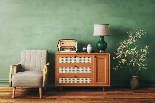 Mid-century modern home decor scene with beige armchair, orange wooden sideboard with cane details, vintage radio, green lamp, and flowering plant against green wall